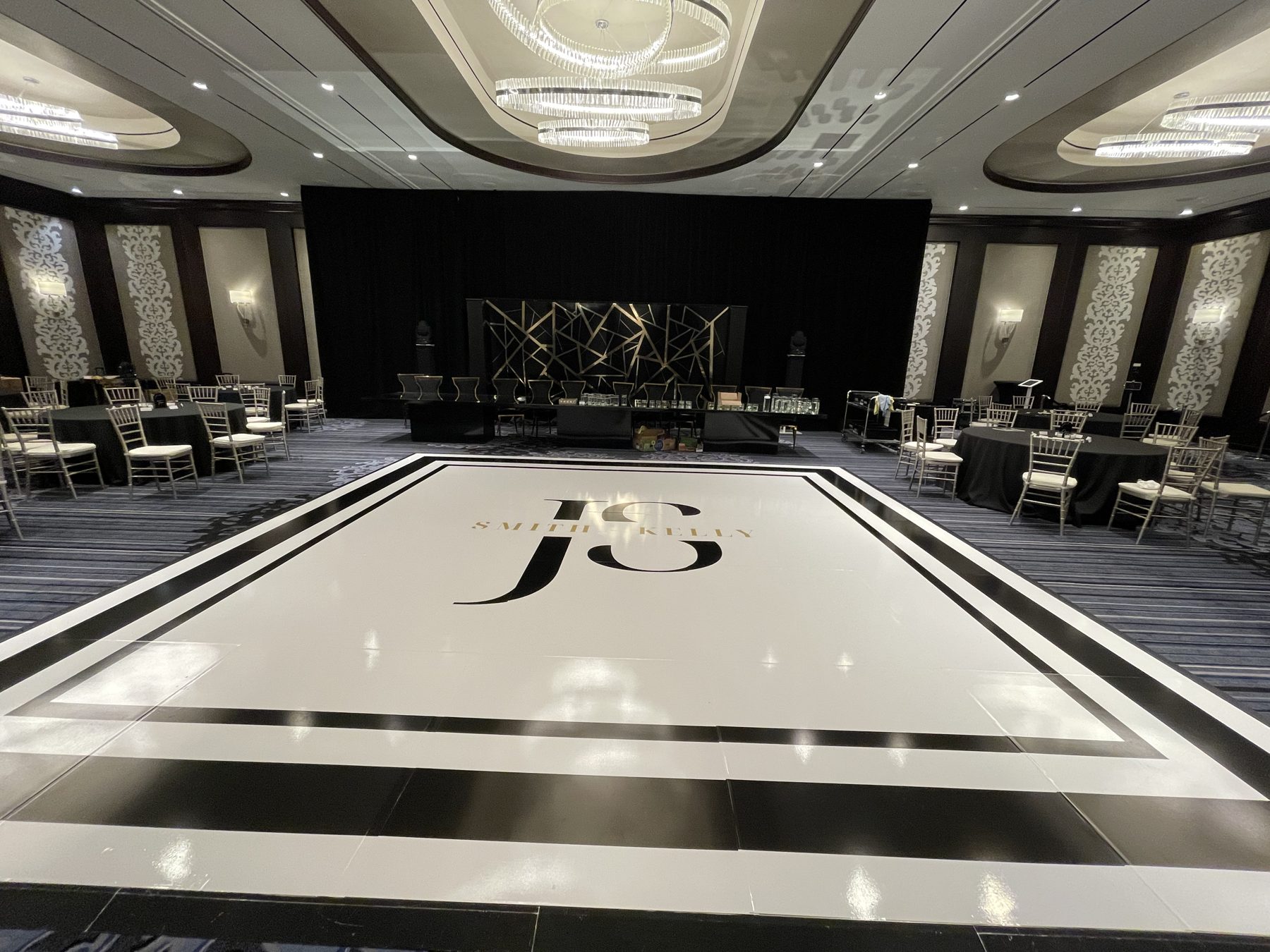 White dance floor wrap with a large black monogram, gold names and a bold double border in a hotel ballroom